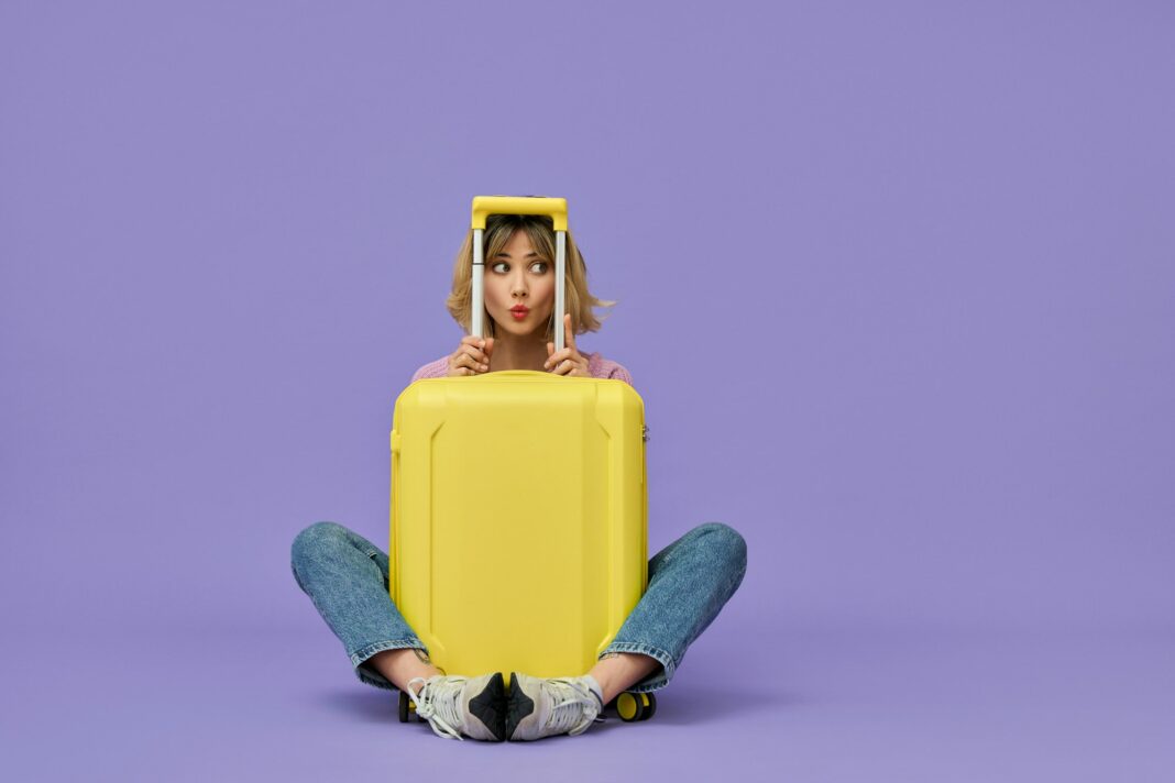 Funny girl traveler sitting with yellow travel suitcase isolated on purple.
